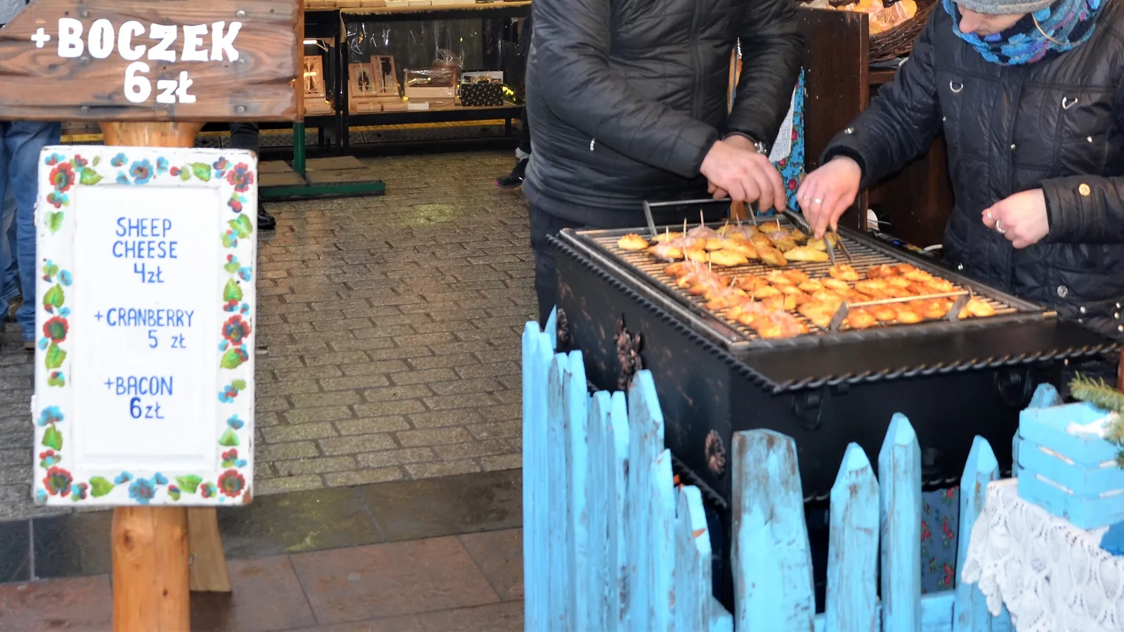Pieces of oscypek cheese on a grill at a market stall, with a painted board offering sheep cheese with cranberry or bacon