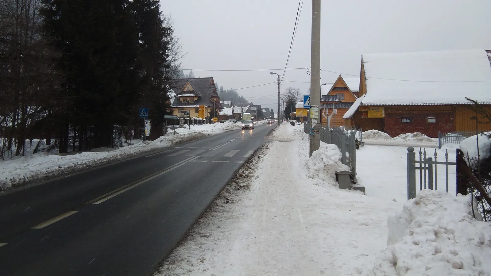 Wet asphalt road between snowbanks and wooden houses in falling snow