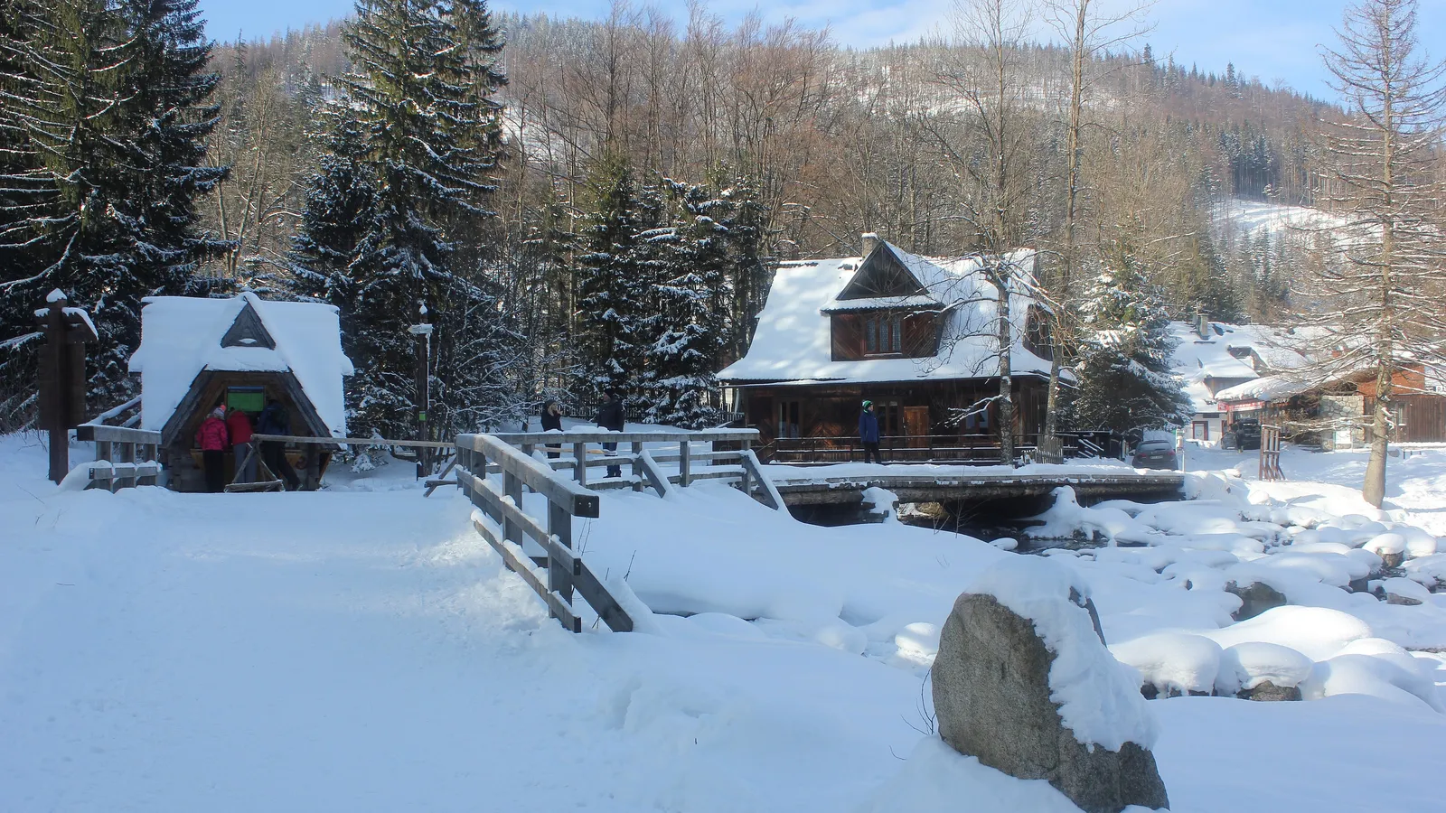 Snow-covered wooden houses and a footbridge over a stream in Kuźnice, with forested slopes behind